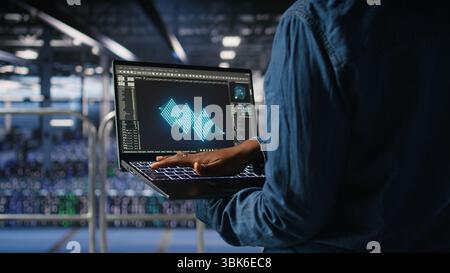 Close up of admin in data center using laptop to monitor neural network LLM visualization. Server hub IT expert overseeing rigs powering machine learning processes using notebook, Stock Photo