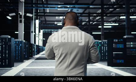 Admin walking in data center, listening music while doing inspection on infrastructure tech. IT specialist in server hub wearing headphones analyzing traffic patterns of equipment mainframes, camera A Stock Photo