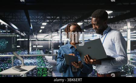 Data center system administrators using laptop to review AI LLM visualizations. African american IT specialists use device to analyze neural networks outputs, ensuring stable gear operations Stock Photo