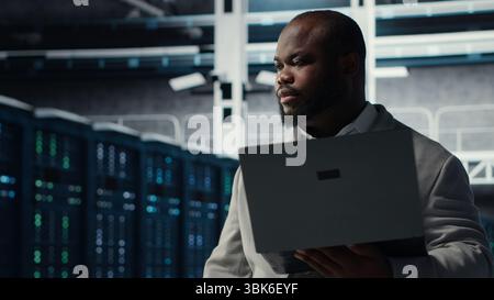 Happy engineer walking in data center, configuring storage devices for minimal latency in AI workloads. Jolly african american worker using device in server farm to build neural networks Stock Photo