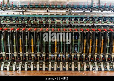A historic textile machine with spindles stands at the Terrade spinning mill (Filature Terrade) on Rue de la Papeterie in Felletin near Aubusson, Creuse, Nouvelle-Aquitaine, France. Bobbins with green, ochre and grey yarn stand in a long row at the bottom. Above them, the threads run through thread guides with red flaps to green rollers and black reels with red levers. The Terrade spinning mill is a family business and one of the last active wool spinning mills and traditional production sites for textile crafts in the country. It was founded in 1923. Managing director Tiffany Terrade runs the Stock Photo