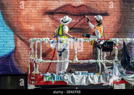 Two artists painting a colorful mural on a brick building while standing on a hydraulic lift. Downtown Calgary, Alberta, Canada. Stock Photo
