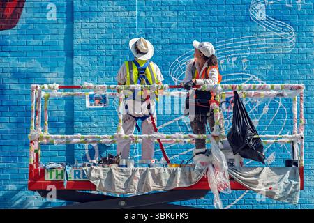 Two artists painting a colorful mural on a brick building while standing on a hydraulic lift. Downtown Calgary, Alberta, Canada. Stock Photo