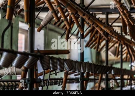 Spindles of a historic textile machine from the 1940s project at an angle from a frame at the Terrade spinning mill (Filature Terrade) on Rue de la Papeterie in Felletin near Aubusson, Creuse, Nouvelle-Aquitaine, France. Some of the brown tubes are wound with light-coloured yarn. A row of grey yarn bobbins runs along a rod in front of them. The Terrade spinning mill is a family business and one of the last active wool spinning mills and traditional production sites for textile crafts in the country. It was founded in 1923. Managing director Tiffany Terrade runs the company in the fifth generat Stock Photo