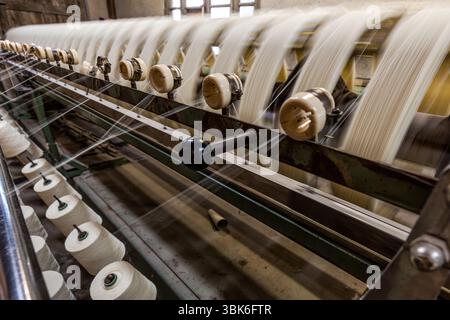An old spinning mill machine at the Terrade spinning mill (Filature Terrade) on Rue de la Papeterie in Felletin near Aubusson, Creuse, Nouvelle-Aquitaine, France, brings together fine yarns. White wool skeins run on rotating swifts, and the threads run down to light-coloured bobbins on green frames. A black knob sits on a lever of the machine. The mill uses largely unchanged machinery and traditional wool processing methods. Most of the mill's mechanical equipment dates from the first half of the 20th century and is designed for durability and precision in traditional craftsmanship. The Terrad Stock Photo