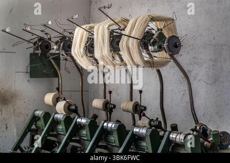 An old spinning mill machine at the Terrade spinning mill (Filature Terrade) winds wool yarn from skeins onto bobbins on Rue de la Papeterie in Felletin near Aubusson, Creuse, Nouvelle-Aquitaine, France. White wool skeins hang on rotating swifts with wire arms. The threads run down to light-coloured bobbins on green frames. Most of the mill's mechanical equipment dates from the first half of the 20th century and is designed for durability and precision in traditional craftsmanship. The Terrade spinning mill is a family business and one of the last active wool spinning mills and traditional pro Stock Photo
