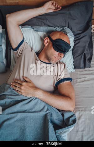 Portrait of middle aged bald man with short beard wearing black sleeping mask lying comfortably in bed during early morning promoting healthy restful Stock Photo