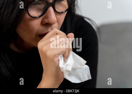 Close-up of an Asian woman wearing glasses coughing into her hand while holding a tissue. Concept of flu, cold, illness, virus infection, or seasonal Stock Photo
