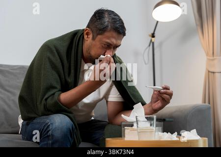 Portrait of a sick Asian man wrapped in a green blanket using tissue and checking thermometer. Concept of cold, flu, fever, and seasonal illness at ho Stock Photo