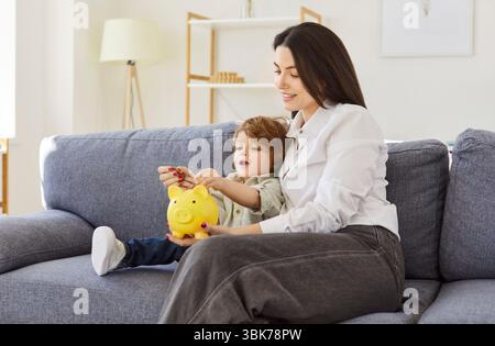 Mother and son, nanny woman sitting together on couch, interacting with yellow piggy bank, learning Stock Photo