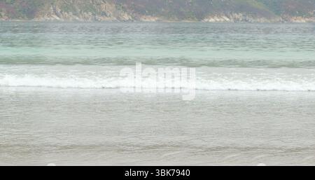 Shallow water waves are rolling onto sandy shore in flat design, overcast sky, rocky hills Stock Photo