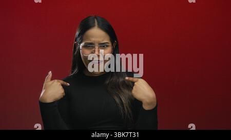 Woman taps finger while gesturing refusal on a red screen, portraying a young latin hispanic model isolated in front of a background wall. Stock Photo