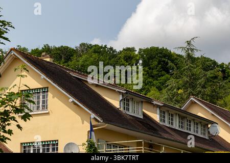 A tan building with a dark roof stands below trees and a cloudy sky, partially framed by foliage on the left in Giverny, France. Stock Photo