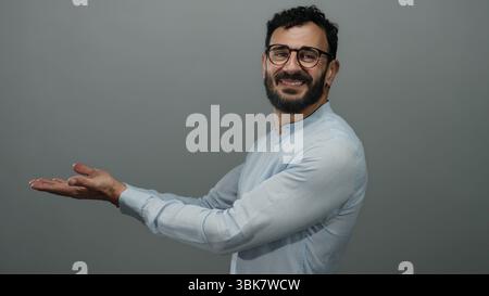 Bearded hispanic man in glasses wearing blue shirt with open palms smiling against grey background, conveying positivity and openness. Stock Photo