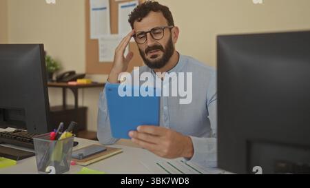 Hispanic man with beard in office looks puzzled holding blue tablet between dual monitors while wearing glasses and light blue shirt indoors. Stock Photo