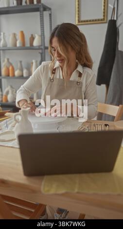 Young blonde woman shapes clay with hands on potters wheel in bright studio space with concentrated expression; creativity focus. Stock Photo