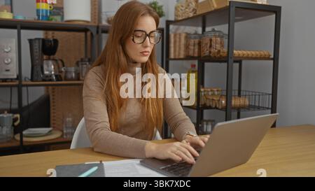 Woman working on laptop in office with redhead hair, wearing glasses, in a professional workplace setting featuring modern interiors and office suppli Stock Photo
