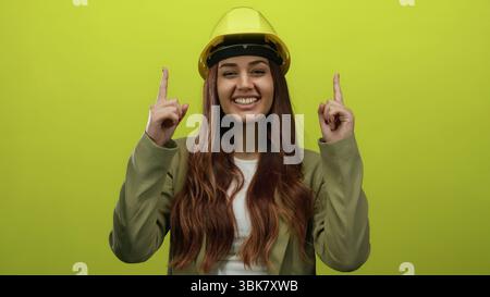 Smiling young hispanic woman architect in hard hat points upward against isolated yellow background, symbolizing construction and engineering creativi Stock Photo