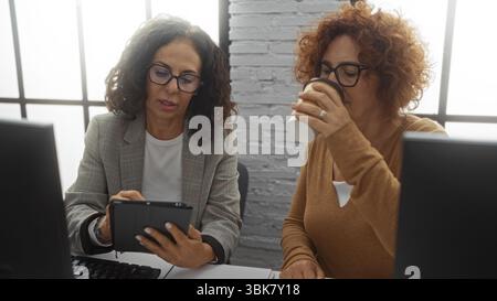 Women working in office with one holding a tablet and the other drinking coffee, suggesting collaboration between business partners in a professional Stock Photo