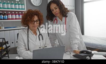 Two hispanic women doctors collaborating in a hospital clinic office, focused on a laptop, surrounded by medical equipment and shelves, depicting a pr Stock Photo