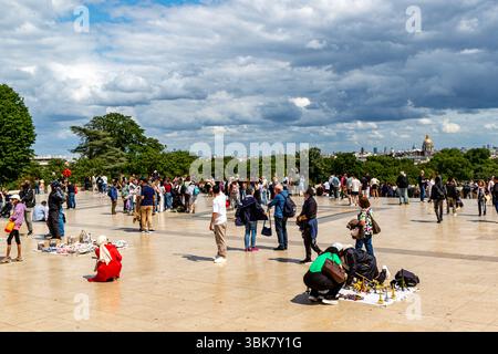 Crowded square in Paris under a cloudy sky, featuring people, trees, distant buildings, and vendors. Stock Photo