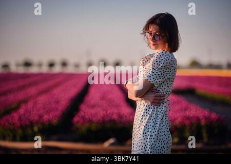 Elegant woman in sunglasses and polka dot dress poses confidently at sunset in a vibrant field of blooming pink tulips. Stock Photo