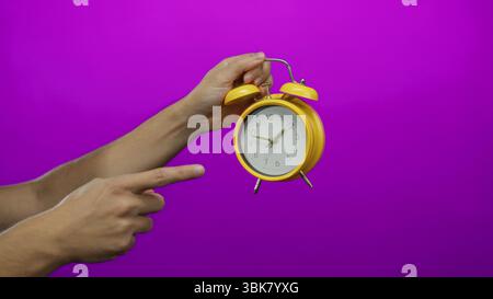 Man holding yellow clock against a vibrant purple wall, with arm pointing, creating a focused and colorful isolated composition. Stock Photo