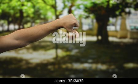 Man's hand giving a thumbs down in a sunlit park, surrounded by lush green trees and blurred background, conveying disapproval or dislike in an outdoo Stock Photo