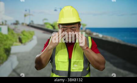 Man resting on seaside promenade wearing construction hat and safety vest looking fatigued under sunny sky near sea possibly taking a break from work Stock Photo