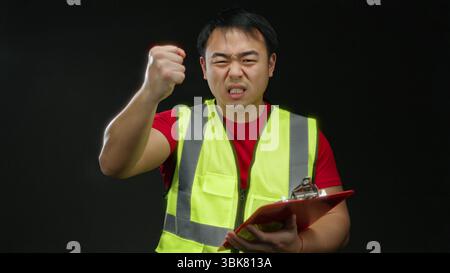 Young asian man wearing safety vest gestures energetically while holding a clipboard against a black background, expressing determination in an isolat Stock Photo