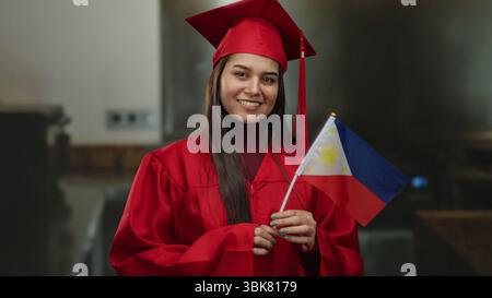 Young woman in a red graduation gown holding a philippines flag smiles proudly indoors, symbolizing academic achievement and cultural pride in a kitch Stock Photo