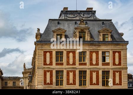 Detailed facade of a large building with a slate roof, multiple windows, and architectural ornamentation against a cloudy sky at Versailles Gardens, P Stock Photo