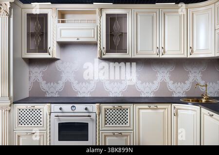 Interior of modern classic style kitchen in beige colors with golden sink basin. Drawers cabinets with lattice and golden ornamental decoration. Dark Stock Photo