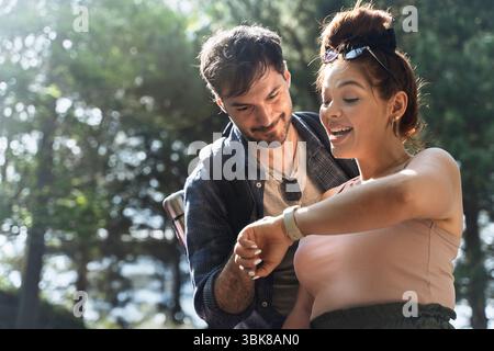 A young couple smiles while looking at a smartwatch on the woman's wrist, checking their progress or fitness goals during a hike in nature Stock Photo