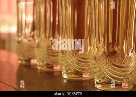 A close-up view of a row of intricately textured glass tumblers placed on a wooden surface. The warm, golden ambient light is reflects. Stock Photo