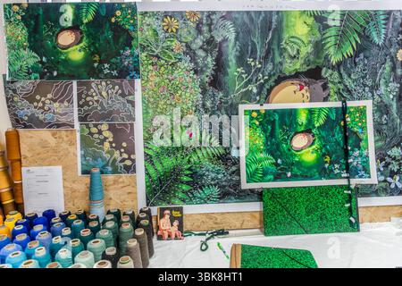 The cartoon and detailed sketches for work on a large-format tapestry featuring the motif from Japanese artist Hayao Miyazaki's "My Neighbour Totoro" hang on a wall of wood-based panels in the creative workshop of the Cité internationale de la tapisserie on Rue des Arts in Aubusson, Creuse, Nouvelle-Aquitaine, France. The large print shows Totoro asleep in the forest among ferns and flowers, with a framed print of the film motif hanging in front of it. A small print of the film motif hangs at the top left, above three motif details with wavy lines, leaves and dots. Cones of yarn in blue, turqu Stock Photo