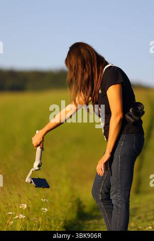 A female photographer with long hair, a camera over her shoulder and a gimbal in her hand, is making a video with her mobile phone. Stock Photo