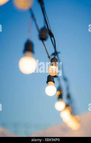 Closeup glowing string bulbs light hanging against blue sky. Creating warm and cozy illumination in event celebration Stock Photo