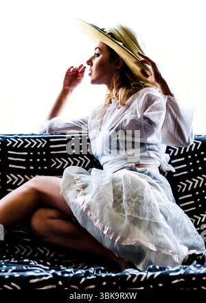 A woman wearing a hat and white clothing is sitting on a couch with a black and white patterned cushion. Stock Photo
