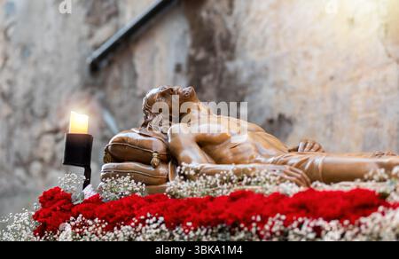 Wooden Statue Of Jesus Christ Resting On A Pillow, Surrounded By Red Carnations And White Flowers, With A Burning Candle Nearby, Capturing Themes Of D Stock Photo