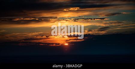 The photo captures a sunset with bright orange and golden sunrays breaking through layered clouds. The horizon is dark, and the sky is filled with dra Stock Photo