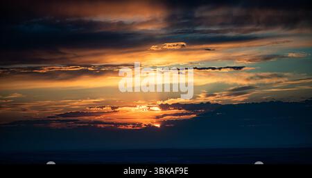 The photo captures a sunset with bright orange and golden sunrays breaking through layered clouds. The horizon is dark, and the sky is filled with dra Stock Photo