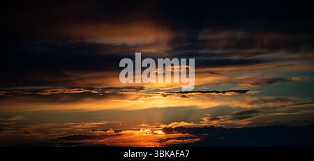 The photo captures a sunset with bright orange and golden sunrays breaking through layered clouds. The horizon is dark, and the sky is filled with dra Stock Photo