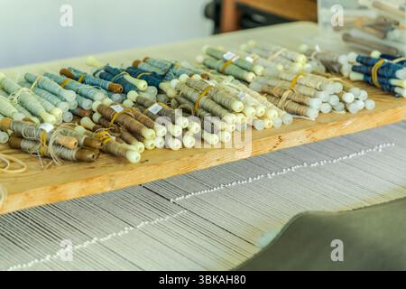 Weaving bobbins (in French flûtes) wound with wool yarn in shades of blue, grey, brown and green lie on a wooden board at the loom at the Robert Four weaving workshop (Manufacture Robert Four) on Rue Madeleine in Aubusson, Creuse, Nouvelle-Aquitaine, France. Small numbered labels mark the colours. The pale warp threads of the loom run underneath. The tapestry "Hommage à George Sand" by Françoise Pétrovitch is being made on this loom. It is made from hand-spun, naturally dyed virgin wool, as is typical for Aubusson tapestries. As part of the "Hommage à George Sand" project, the weaving of a lar Stock Photo