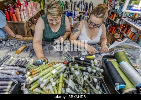 Weavers Frédérique (left) and Agathe (right) work with lowered eyes at the low-warp loom of the Robert Four weaving workshop (Manufacture Robert Four) on Rue Madeleine in Aubusson, Creuse, Nouvelle-Aquitaine, France. They are surrounded by wool yarns and weaving bobbins (in French flûtes) in many shades of colour for the tapestry "Hommage à George Sand" by French artist Françoise Pétrovitch. Baskets of bobbins wound with green, violet and grey wool stand on the warp threads. The older weaver wears glasses and a green lace top, the younger one glasses, a white vest top and a flower tattoo on he Stock Photo