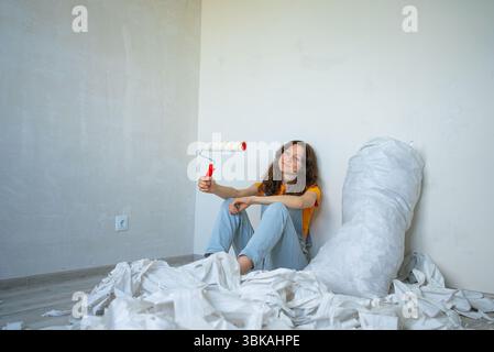 Young woman taking a break from removing old wallpaper in a room, holding a paint roller and sitting on the floor among the scraps, ready for the next step of home renovation Stock Photo