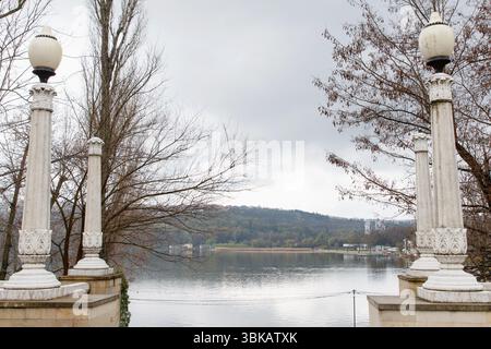 Four white ornate stone lamp columns frame a scenic lakeside view with forested hills, under an overcast gray sky, symbolizing tranquility Stock Photo