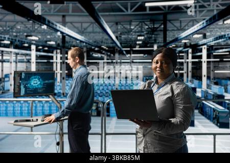 Portrait of smiling female engineer in server farm next to colleague doing infrastructure AI automatization. Happy african american woman in data center doing maintenance using artificial intelligence Stock Photo