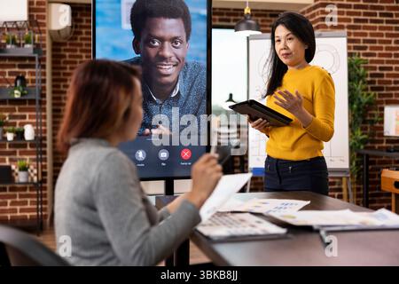 Businesswomen collaborating on startup project, utilizing led screen for a video call with an african american client. Female coworkers in virtual meeting with male manager, providing company updates. Stock Photo