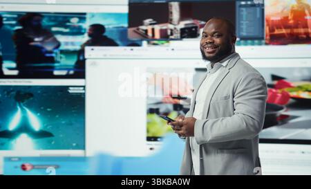 Portrait of smiling man browsing social networks feed on smartphone in front of videowall. Jolly african american person uses phone, floating icons highlighting interactive engagement online, camera A Stock Photo